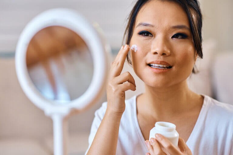 Beautiful Asian woman applying face cream while looking herself in a mirror